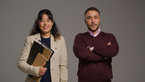 Retrato-De-Estudio-De-Profesores-Hombres-Y-Mujeres-Sonrientes-Sosteniendo-Una-Carpeta-Y-Cruzando-Los-Brazos-Sobre-Un-Fondo-Gris