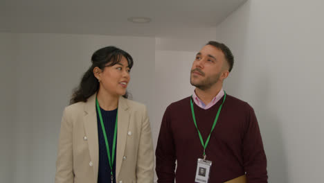 Male-And-Female-Teachers-Wearing-Security-Lanyards-Talking-As-They-Walk-Along-Corridor-In-School-Building-Together-1