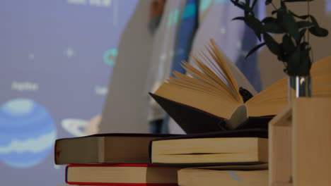 Close-Up-Of-Textbooks-On-Desk-As-Teacher-Gives-Maths-Or-Science-Lesson-In-School-Standing-At-The-Front-Of-Class-With-Projection-Of-Graph