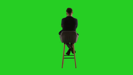 Rear-View-Studio-Shot-Of-Man-Sitting-On-Stool-Facing-Away-From-Camera-Against-Green-Screen