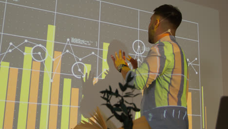 Male-Teacher-Giving-Maths-Or-Science-Lesson-In-School-Standing-At-The-Front-Of-Class-With-Projection-Of-Graph