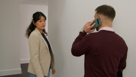 Male-Teacher-Talking-On-Mobile-Phone-Passing-Female-Colleague-As-They-Walk-Along-Corridor-In-School-Building