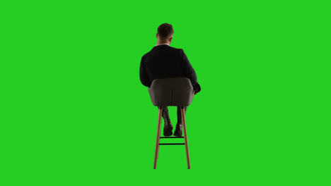 Rear-View-Studio-Shot-Of-Man-Sitting-On-Stool-Facing-Away-From-Camera-Against-Green-Screen