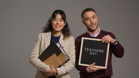 Retrato-De-Estudio-De-Profesores-Hombres-Y-Mujeres-Sonrientes-Sosteniendo-Un-Tablón-De-Anuncios-Leyendo-El-Día-Del-Maestro-Contra-Un-Fondo-Gris