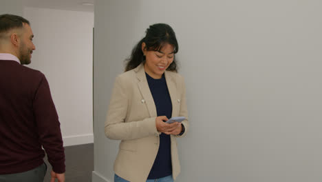 Female-Teacher-Using-Mobile-Phone-Passing-Male-Colleague-As-They-Walk-Along-Corridor-In-School-Building