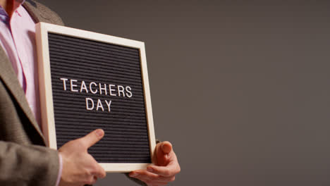 Close-Up-Studio-Shot-Of-Male-Teacher-Standing-Against-Grey-Background-Holding-Notice-Board-Reading-Teachers-Day