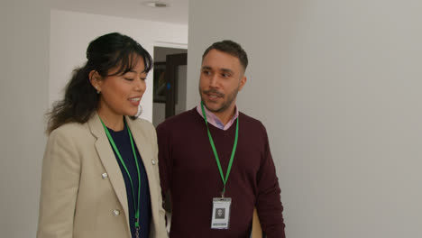 Male-And-Female-Teachers-Wearing-Security-Lanyards-Talking-As-They-Walk-Along-Corridor-In-School-Building-Together