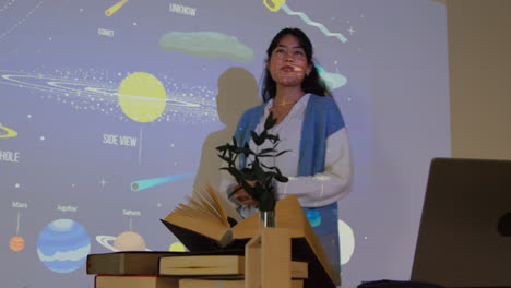 Female-Teacher-Giving-Astronomy-Lesson-In-School-Standing-At-The-Front-Of-Class-With-Projection-Of-The-Solar-System