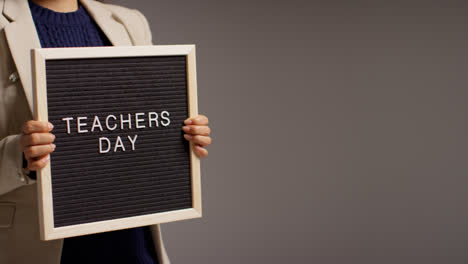 Close-Up-Studio-Shot-Of-Female-Teacher-Standing-Against-Grey-Background-Holding-Notice-Board-Reading-Teachers-Day-1