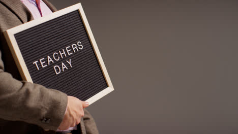 Close-Up-Studio-Shot-Of-Male-Teacher-Standing-Against-Grey-Background-Holding-Notice-Board-Reading-Teachers-Day-1