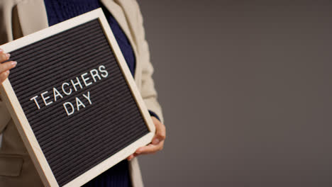 Close-Up-Studio-Shot-Of-Female-Teacher-Standing-Against-Grey-Background-Holding-Notice-Board-Reading-Teachers-Day