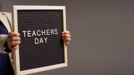 Close-Up-Studio-Shot-Of-Female-Teacher-Standing-Against-Grey-Background-Holding-Notice-Board-Reading-Teachers-Day-2