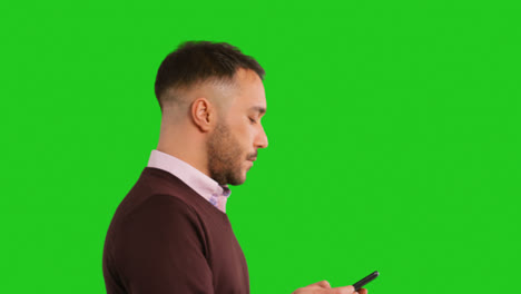 Close-Up-Studio-Shot-Of-Walking-Businessman-Using-Mobile-Phone-To-Message-Or-Browse-Online-Standing-Against-Green-Screen