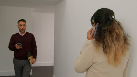 Female-Teacher-Talking-On-Mobile-Phone-Passing-Male-Colleague-As-They-Walk-Along-Corridor-In-School-Building