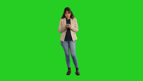 Retrato-De-Estudio-De-Cuerpo-Entero-De-Una-Maestra-O-Empresaria-Parada-Frente-A-Una-Pantalla-Verde-Usando-Un-Teléfono-Móvil-Para-Enviar-Mensajes-O-Navegar-En-Línea.