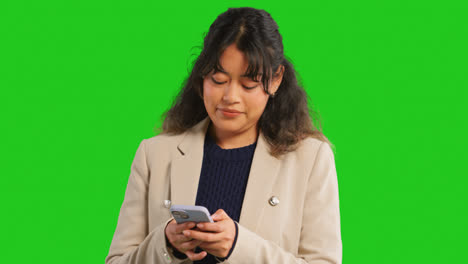 Close-Up-Studio-Shot-Of-Smiling-Businesswoman-Using-Mobile-Phone-To-Message-Or-Browse-Online-Standing-Against-Green-Screen