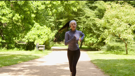 Front-View-Of-Young-Woman-Exercising-Running-Through-City-Park-Wearing-Wireless-Earbuds