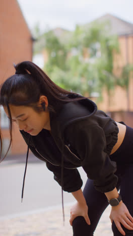 Video-Vertical-De-Una-Mujer-Joven-Haciendo-Ejercicio-Corriendo-En-Una-Calle-Urbana-Usando-Auriculares-Inalámbricos-Y-Mirando-Un-Reloj-Inteligente