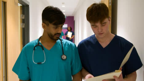 Two-Medical-Staff-Discuss-Chart-Walking-In-Corridor