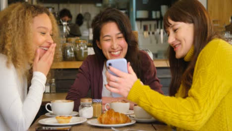 Pan-of-Woman-Showing-Her-Friends-Something-Funny-On-Phone-In-Cafe
