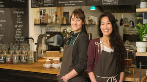 Portrait-of-two-female-baristas-at-a-cafe-smiling-to-camera