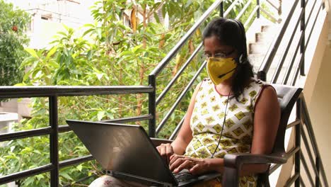 Nahaufnahme-Einer-Indischen-Frau-Mit-Sicherheitsmaske-Und-Headset-Mikrofon,-Die-Mit-Einem-Laptop-Arbeitet,-Der-Während-Der-Covid19-Coronavirus-Pandemie-Auf-Dem-Balkon-Sitzt