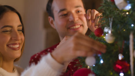 Tracking-Shot-From-Behind-Christmas-Tree-As-Young-Couple-Decorate-It