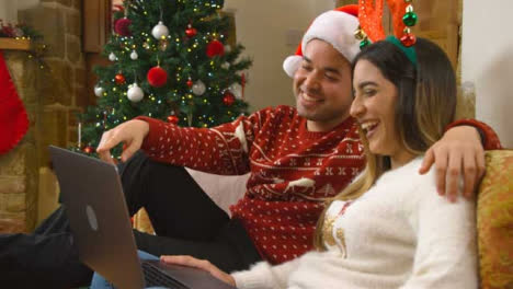 Mittlere-Aufnahme-Eines-Jungen-Paares,-Das-Während-Eines-Weihnachtsvideoanrufs-Lacht-Und-Mit-Der-Laptopkamera-Spricht
