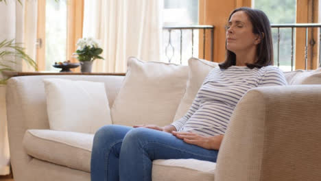 Pull-Focus-Shot-of-Middle-Aged-Woman-Meditating-On-Sofa