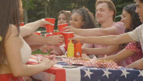 Sliding-Shot-of-Group-of-American-Friends-Sitting-Around-Table-Bringing-Their-Drinks-Together