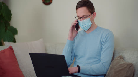 Man-Wearing-Protective-Mask-While-Working-On-Laptop-And-Talking-On-Mobile-Phone