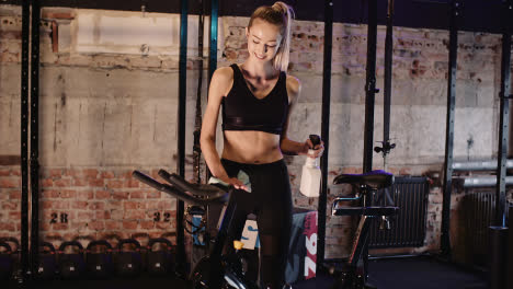 Smiling-Confident-Female-Instructor-Cleaning-Handle-Of-Stationary-Bike-At-Fitness-Club