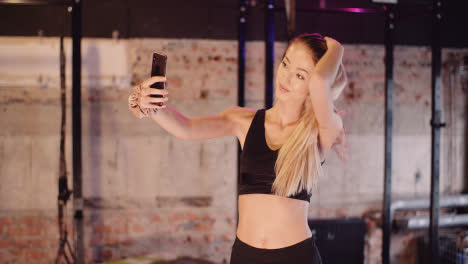 Beautiful-Female-Athlete-Adjusting-Hair-While-Taking-Selfie-Through-Smartphone-At-Health-Studio