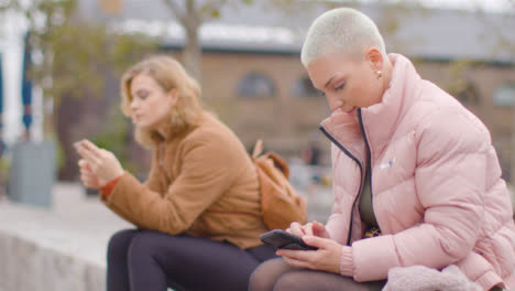 Medium-Shot-of-Two-Young-Women-Sitting-Outside-On-Their-Phones