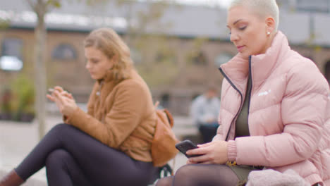 Tracking-Shot-of-Two-Young-Women-Sitting-Outside-On-Their-Phones