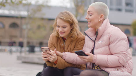 Tracking-Shot-of-Two-Female-Friends-Sitting-Outside-On-Their-Phones