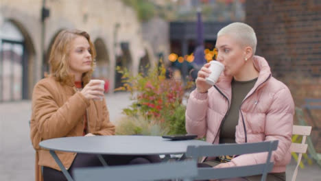 Wide-Shot-of-Two-Young-Women-Sitting-at-Outdoor-Table-Drinking-Coffee