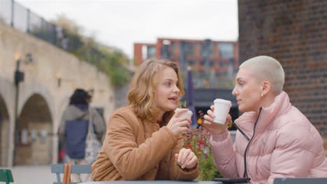 Low-Angle-Shot-of-Two-Young-Women-Sitting-at-Outdoor-Table-Drinking-Cups-of-Coffee