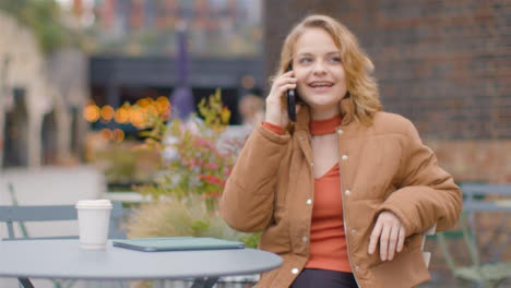 Medium-Shot-of-Young-Woman-Sitting-at-Outdoor-Table-Answering-Phone-Call
