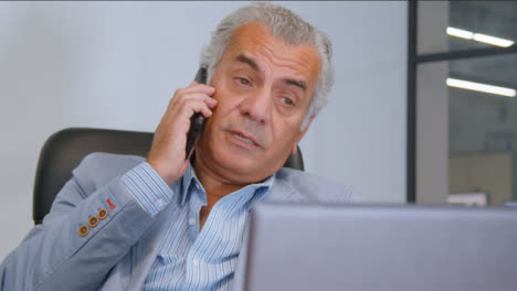 Close-Up-Shot-of-a-Senior-Man-Sitting-at-His-Desk-and-Talking-On-the-Phone