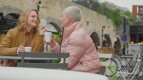 Med-Shot-of-Two-Young-Women-Sitting-Outside-Talking-and-Drinking-Coffee