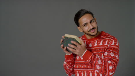 Portrait-Shot-of-Young-Man-In-Christmas-Sweater-Shaking-Gift-with-Copy-Space