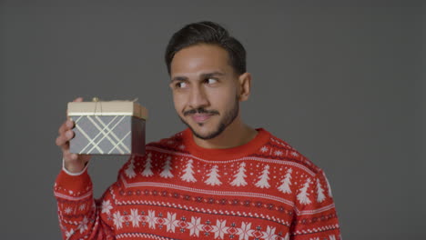 Portrait-Shot-of-Young-Man-In-Christmas-Sweater-Shaking-Gift