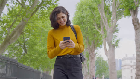 Toma-De-Seguimiento-En-ángulo-Bajo-De-Una-Joven-Estudiante-Caminando-Mientras-Mira-Su-Teléfono-Inteligente