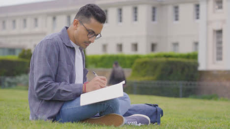 Tiro-Medio-De-Un-Estudiante-Masculino-Usando-El-Bloc-De-Notas-En-El-Parque-Universitario