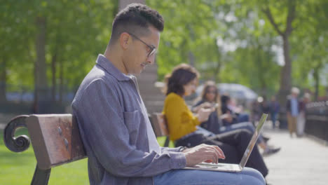 Plano-Medio-Del-Hombre-Usando-Una-Computadora-Portátil-Sentado-En-Un-Banco-Del-Parque