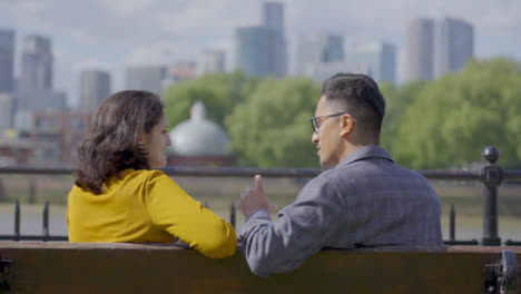 Mid-Shot-of-Two-Friends-Sitting-and-Talking-on-Bench-Against-City-Skyline