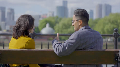 Mid-Shot-of-Two-Friends-Sit-and-Talk-on-Bench-Against-London-City-Skyline