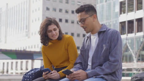 Arc-Shot-of-Two-Students-Talking-and-Using-Phones-with-City-Backdrop