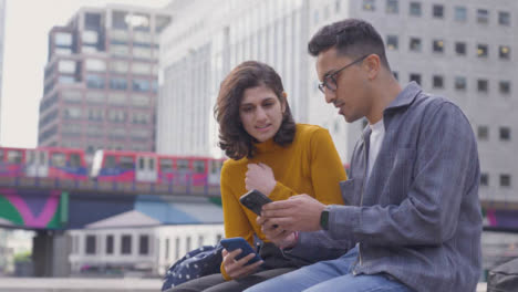 Arc-Shot-of-Two-Students-Talking-and-Using-Smartphone-with-City-Backdrop